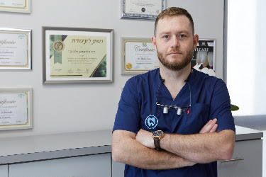 ד"ר אלכסנדר גולדובסקי במרפאת סיטיסמייל באשדוד, על רקע קיר התעודות של המרפאה